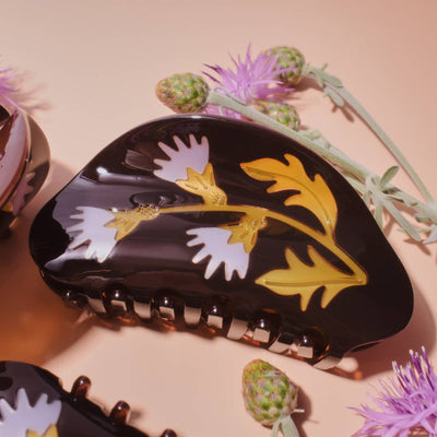 
                    
                      Milk Thistle hair claw, glossy black cellulose acetate with white and yellow floral pattern, close-up flat lay with thistle…
                    
                  