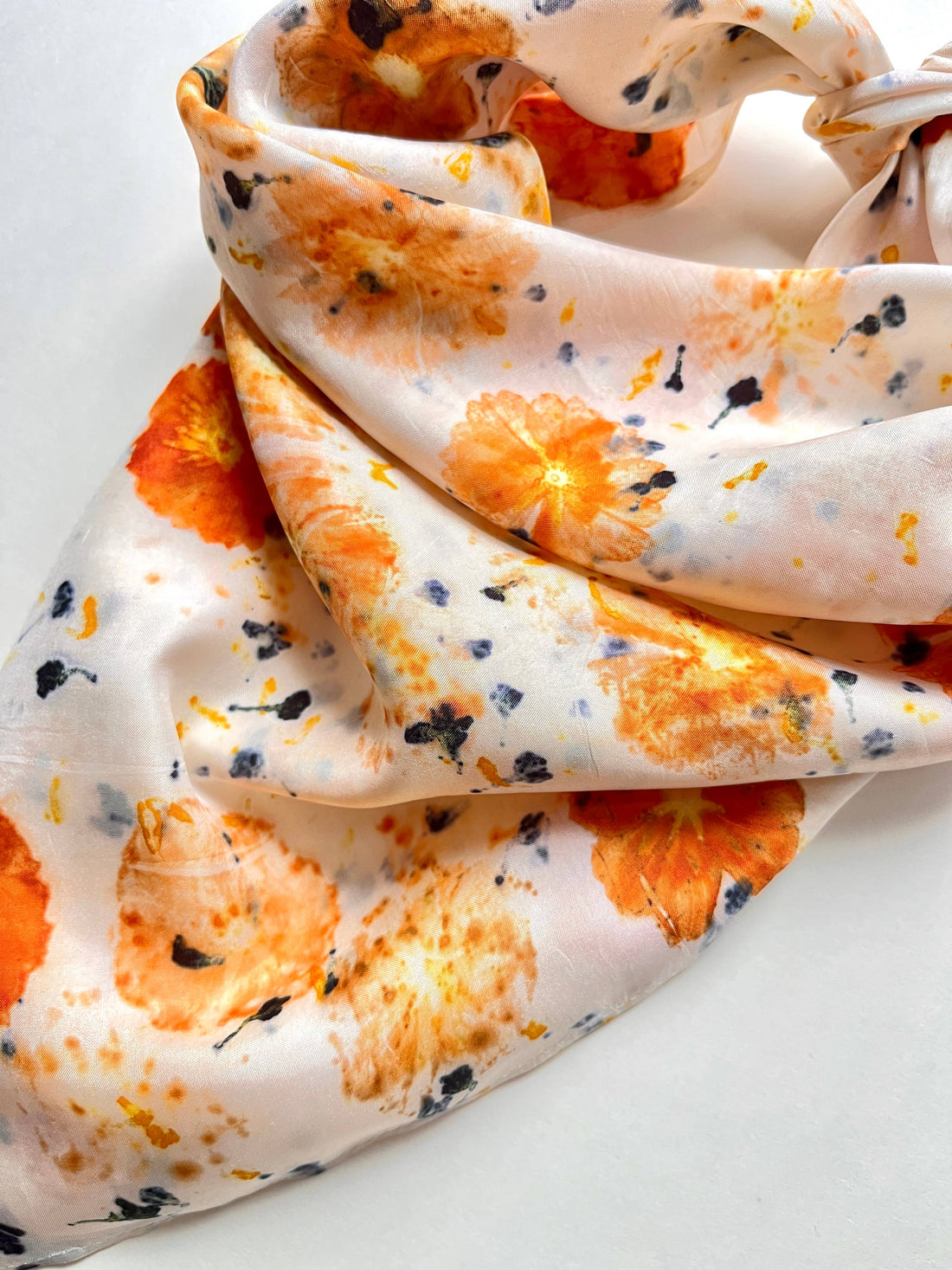 Abby silk habotai bandana scarf, draped close-up showing vibrant orange floral imprints and dark blue specks on creamy…
