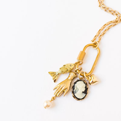 
                    
                      Carabiner Charm Necklace. Close-up of gold-plated carabiner clasp, detailed fish, hand, heart, and black & white cameo…
                    
                  