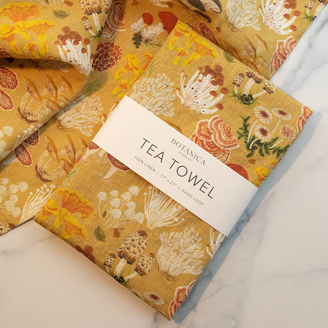 Botanica Paper Co. Mushrooms 100% linen tea towel, mustard yellow with colorful mushroom print, folded with brand band…