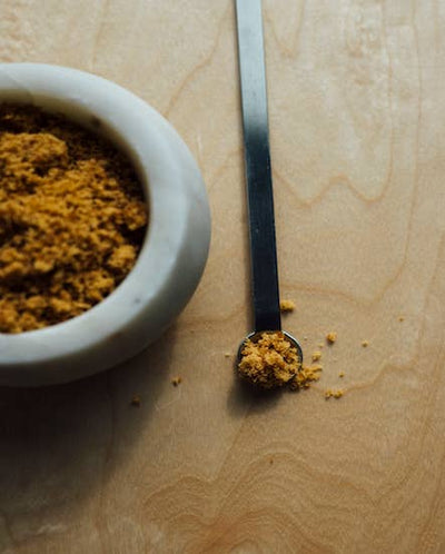 
                    
                      Artisanal Jaggery sweetener, golden-brown granular, a spoonful on a silver measuring spoon next to a white bowl.
                    
                  
