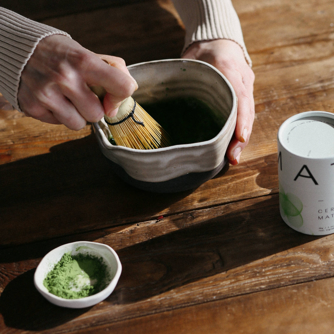 Hands whisking Dona Ceremonial Matcha in a textured ceramic bowl, with loose green powder and white canister on a wooden…