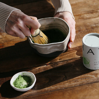 
                    
                      Hands whisking Dona Ceremonial Matcha in a textured ceramic bowl, with loose green powder and white canister on a wooden…
                    
                  