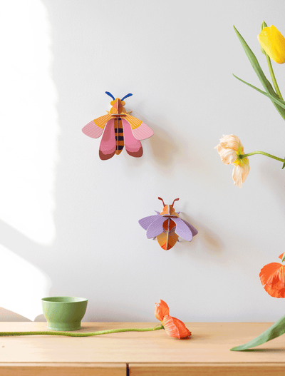 
                    
                      Pink Bee 3D build kit, pink, yellow, and blue recycled cardboard, displayed on a white wall beside a purple bee kit.
                    
                  