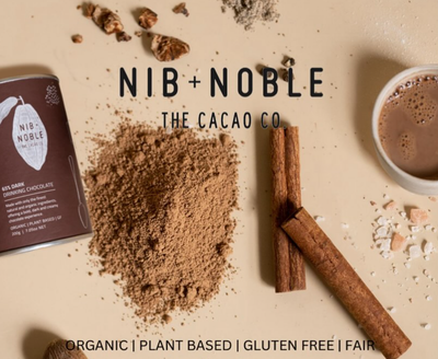 
                    
                      Nib + Noble Organic Spiced Chai Powder flat lay: brown powder, cinnamon sticks, cocoa nibs, and '65% Dark Drinking…
                    
                  