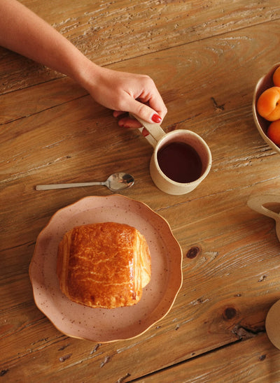 
                    
                      Pink handcrafted stoneware plate with scalloped edge, styled with a pastry, overhead view on a wooden table.
                    
                  
