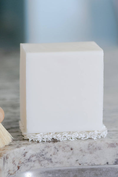 
                    
                      No Tox Life Dish Block solid white soap on white scrub pad, alongside wooden brush, close-up on granite countertop.
                    
                  