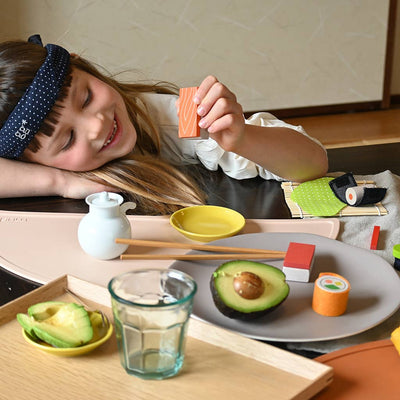 
                    
                      Smiling child playing with Sushi Party Set toys, holding an orange wooden salmon piece, with beechwood sushi rolls, avocado,…
                    
                  