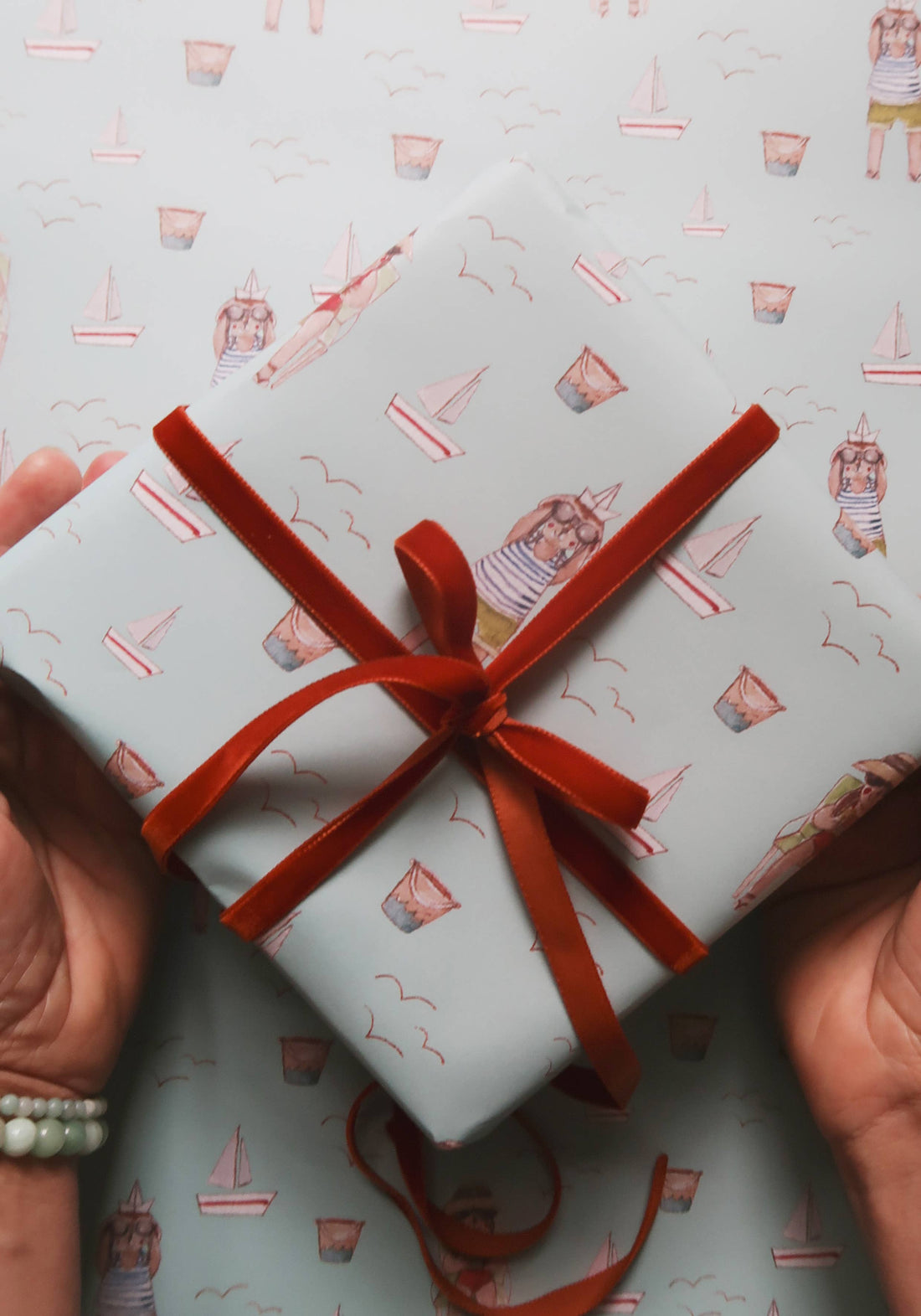 Agnese Grīnberga Summer Wrapping Paper, light blue with watercolor sailboats and beach-goers, tied with red velvet ribbon,…