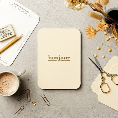 
                    
                      Katie Leamon 'bonjour' Cloth-Covered Postcard, cream cotton with gold foil text, flat lay showing postcard collection and…
                    
                  
