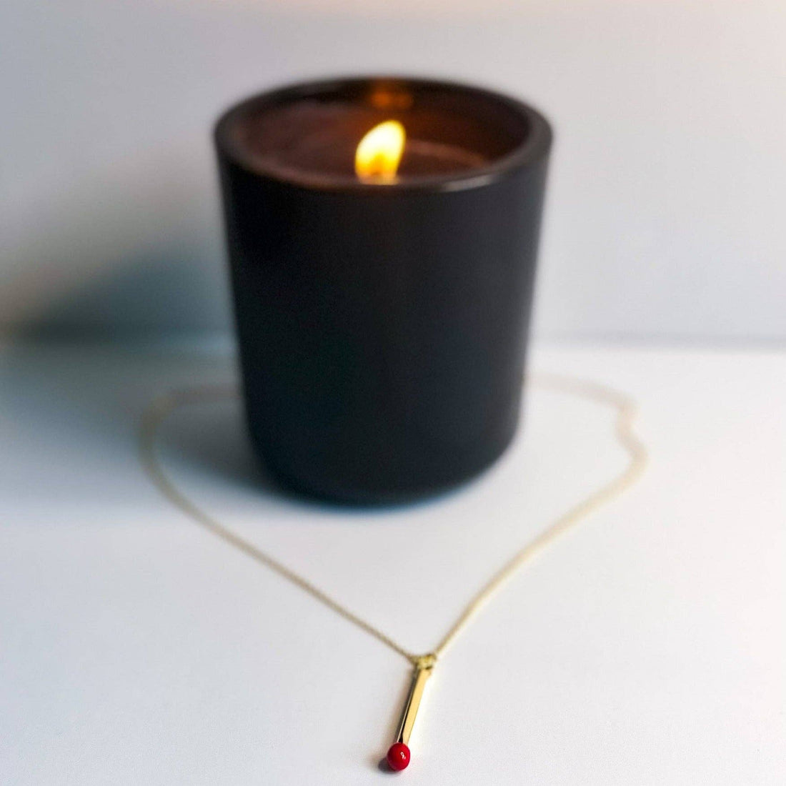 Matchstick Necklace, gold-plated with red tip, displayed flat on a white surface, with a softly blurred lit candle in the…