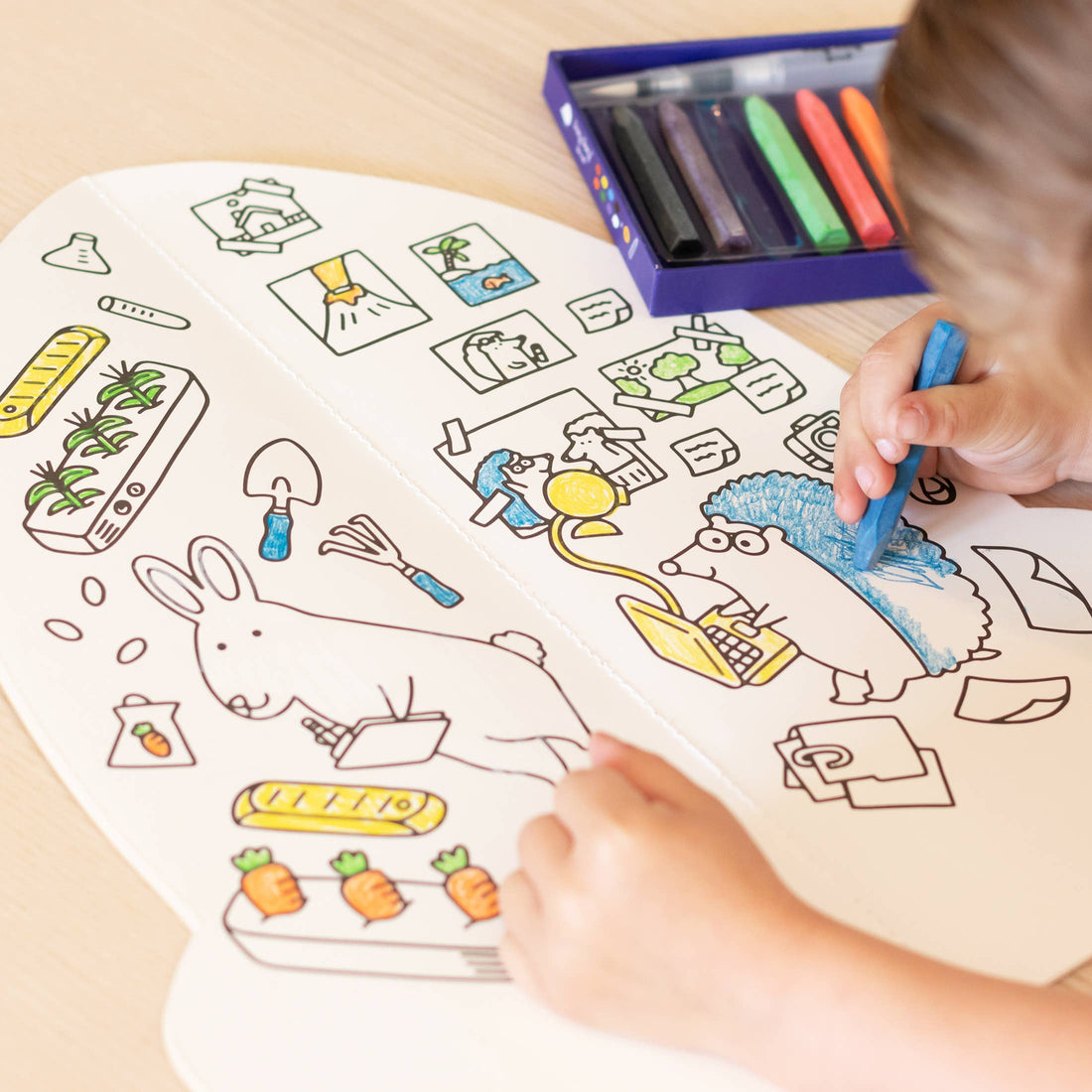 Jumbo Rocket paper coloring book, child coloring a hedgehog blue with a crayon, open spread with various illustrations on…