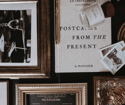 
                    
                      Postcards from the Present travel journal, premium linen cover, flat lay amidst vintage frames, gold pen, and travel photos.
                    
                  