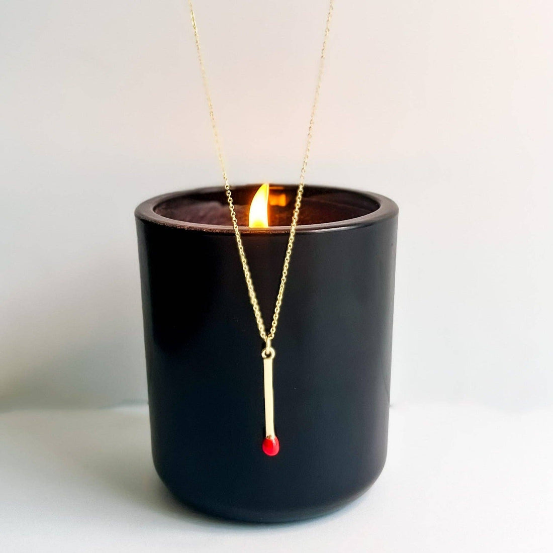 Matchstick necklace, gold-plated with red tip, dangling above a lit candle in a black vessel.
