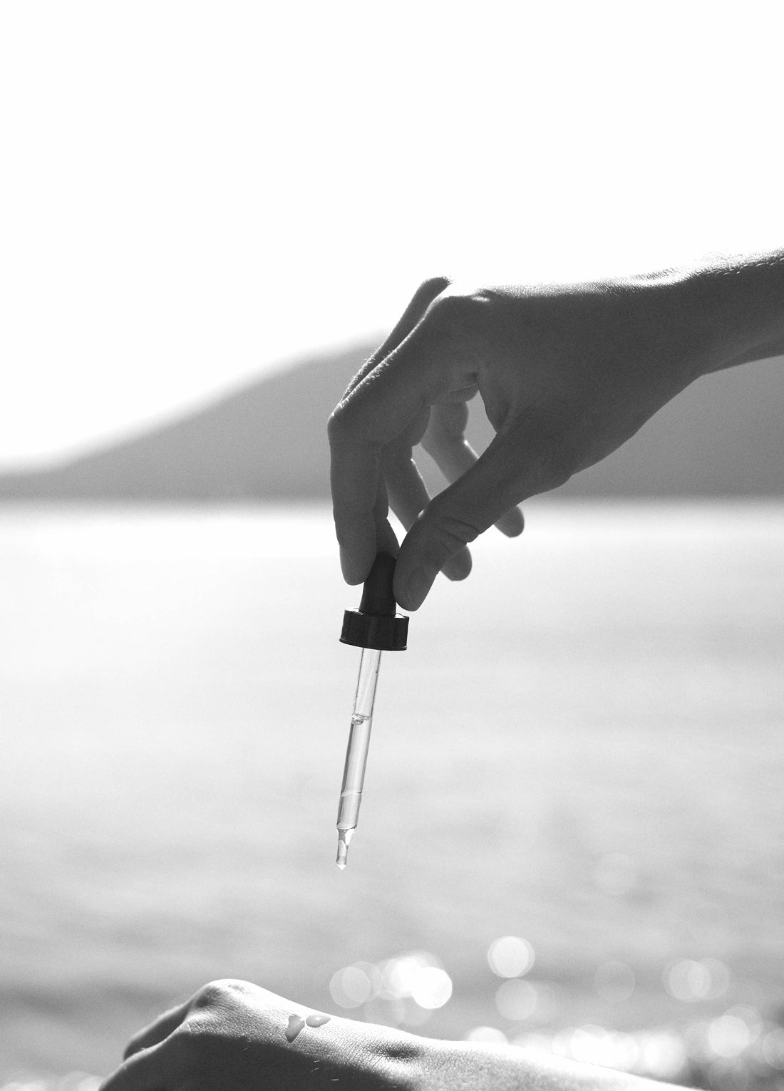 Shave Oil, clear glass dropper with dark cap dispensing oil drops onto skin, blurred water background.