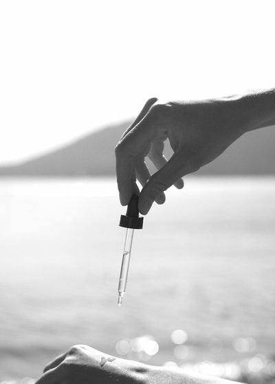 
                    
                      Shave Oil, clear glass dropper with dark cap dispensing oil drops onto skin, blurred water background.
                    
                  