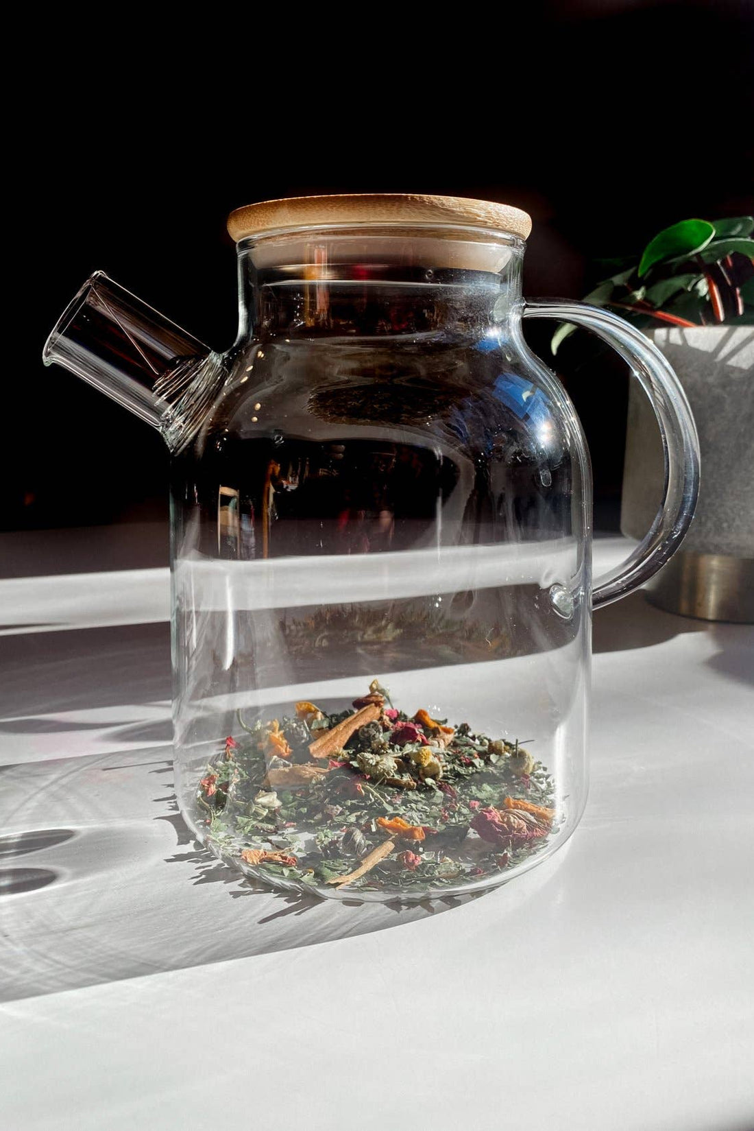 Heat-Resistant Glass Teapot with Bamboo Lid, filled with colorful loose leaf tea, detailed angled view on sunlit surface.
