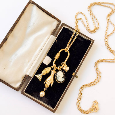 
                    
                      Close-up of Gold Carabiner Charm Necklace with varied charms and pearl, resting in an open velvet box.
                    
                  
