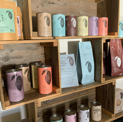 
                    
                      Nib + Noble Organic Spiced Chai Powder, light brown kraft pouch with teal label, displayed on a rustic wooden shelf.
                    
                  