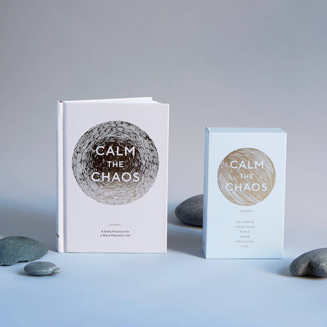 Calm The Chaos mindfulness card deck (light blue box) and companion journal (white), upright with gold design & grey stones.