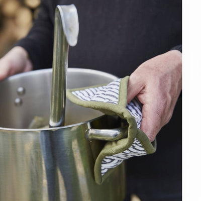 
                    
                      Aux Petits Oignons oven mitt with green trim and striped textile, worn on hand holding a stainless steel pot handle.
                    
                  