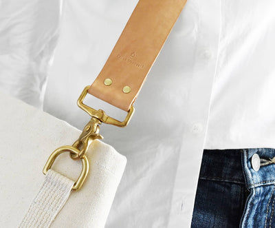 
                    
                      Celina Mancurti Everyday Tote Bag, tan leather strap with embossed brand, raw brass hardware attached to natural canvas,…
                    
                  