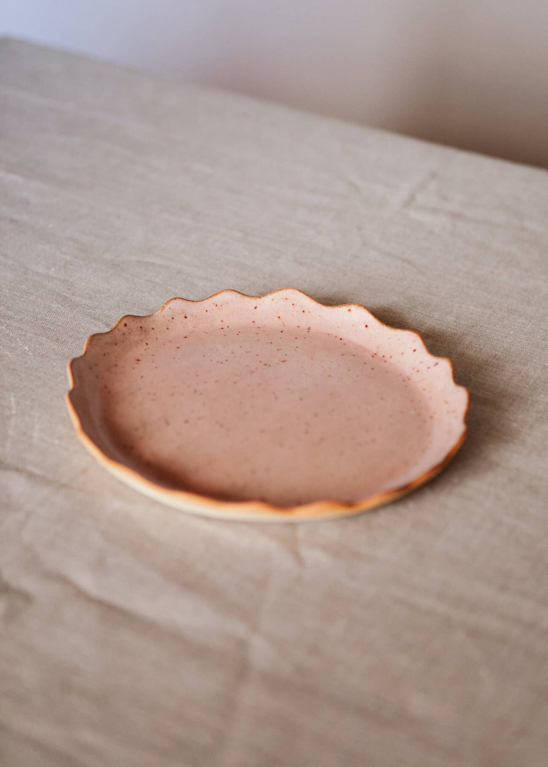 Handcrafted Stoneware Plate, speckled terracotta pink glaze, raw scalloped edge, angled close-up view on textured beige…