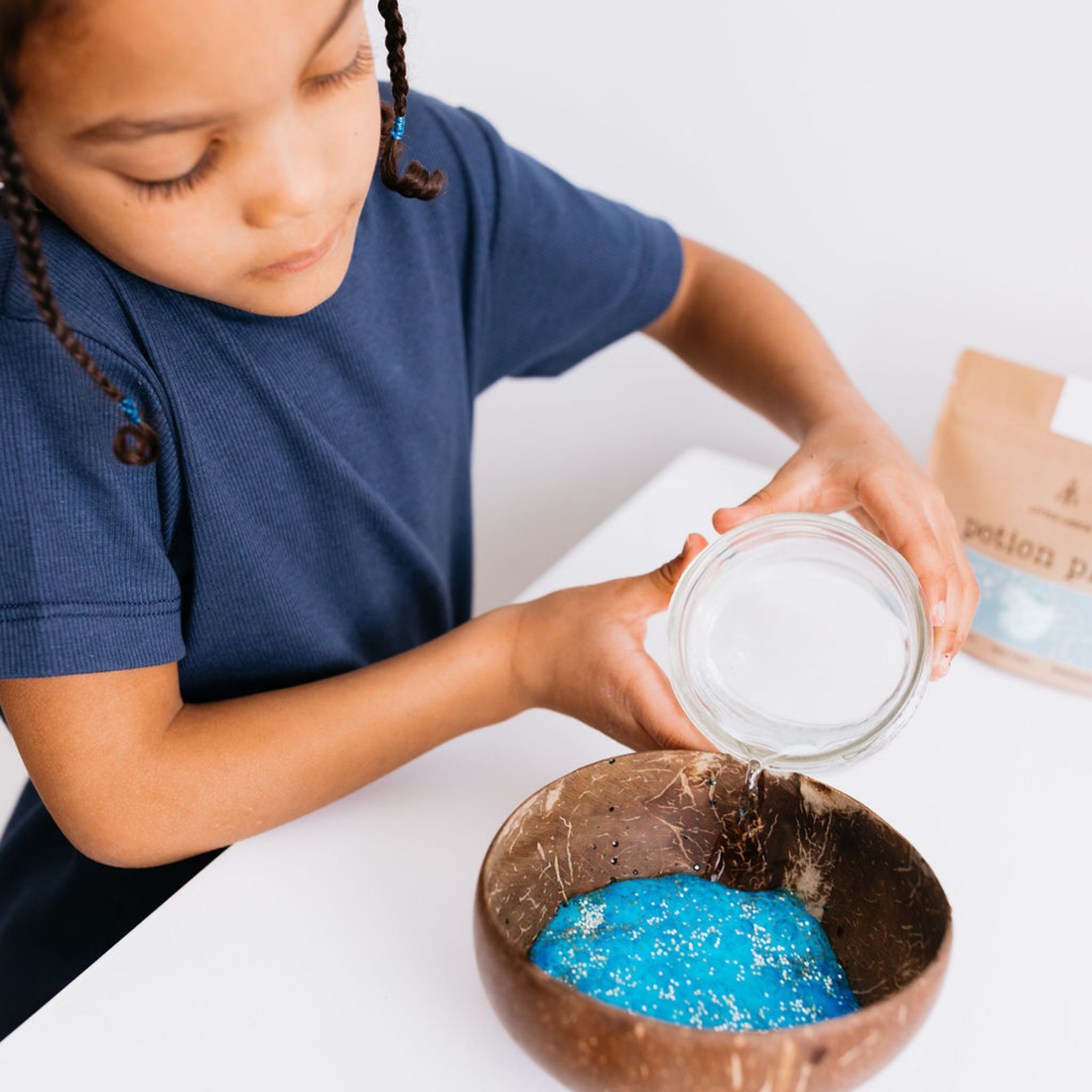 Bravery Spell sensory play kit: child pouring water into blue glitter powder in a natural coconut shell bowl.