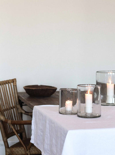 
                    
                      Multiple White Pillar Candles, lit in clear glass hurricane lamps, arranged on a white tablecloth.
                    
                  