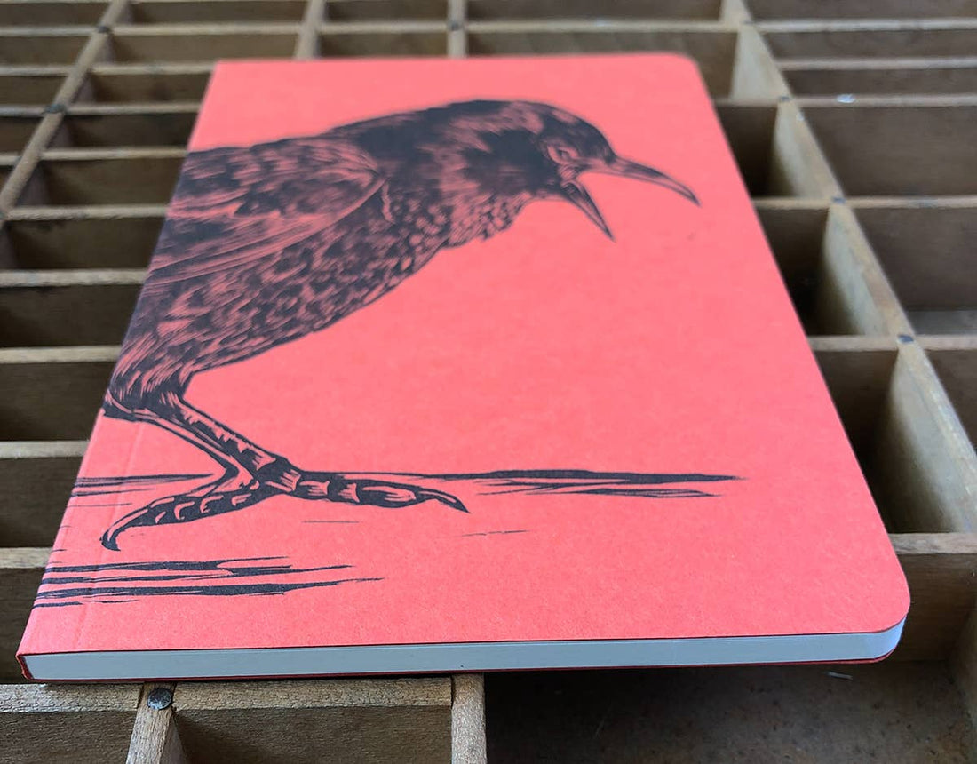 Crow Large Letterpress notebook, red-orange recycled paper cover with black crow illustration, angled view on a wooden type…