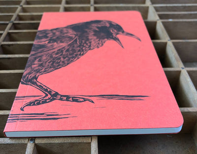 
                    
                      Crow Large Letterpress notebook, red-orange recycled paper cover with black crow illustration, angled view on a wooden type…
                    
                  