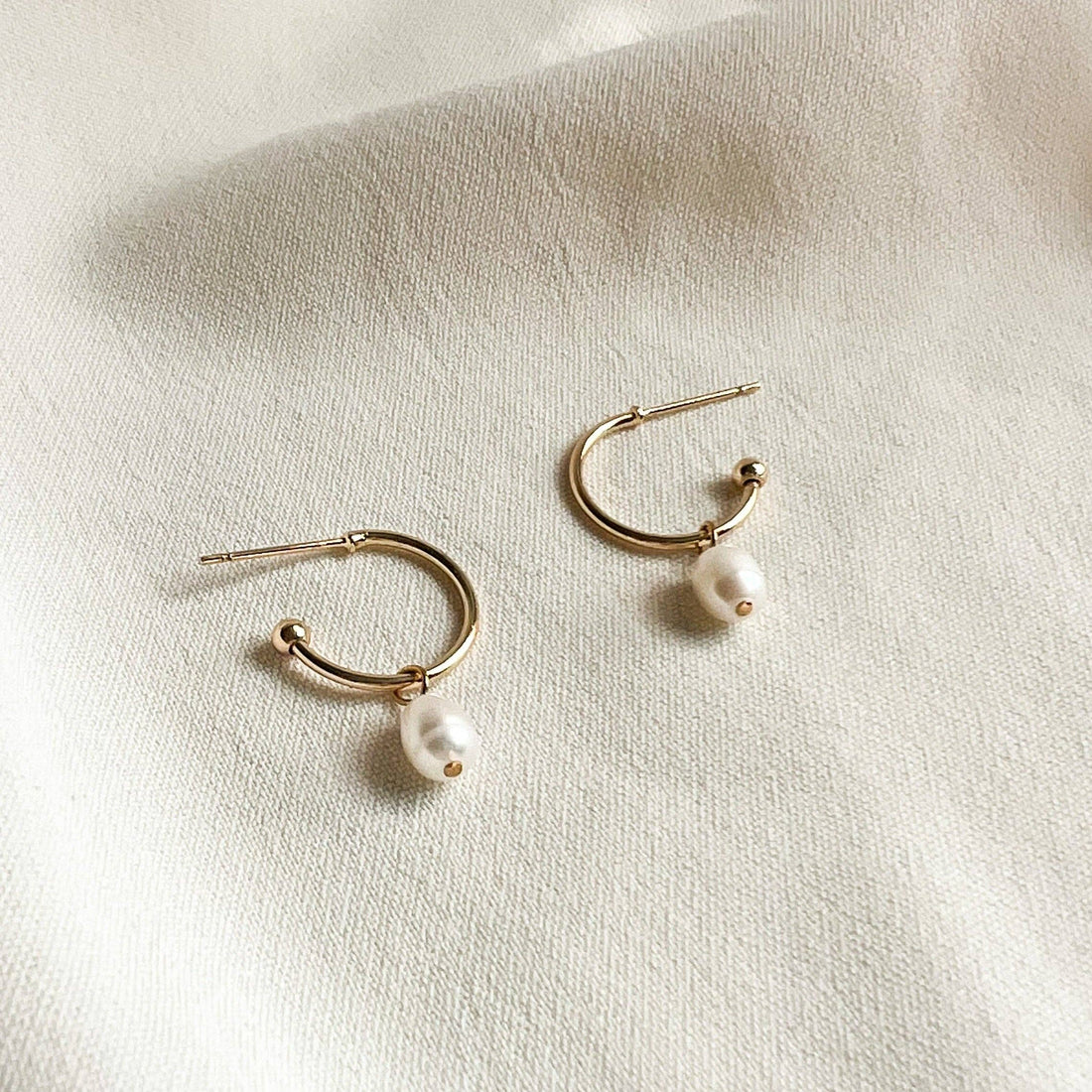 Natalie Clare Chloe Pearl hoop earrings, 18K gold-plated brass with white pearls, flat lay pair on textured fabric.