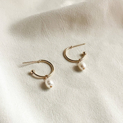 
                    
                      Natalie Clare Chloe Pearl hoop earrings, 18K gold-plated brass with white pearls, flat lay pair on textured fabric.
                    
                  