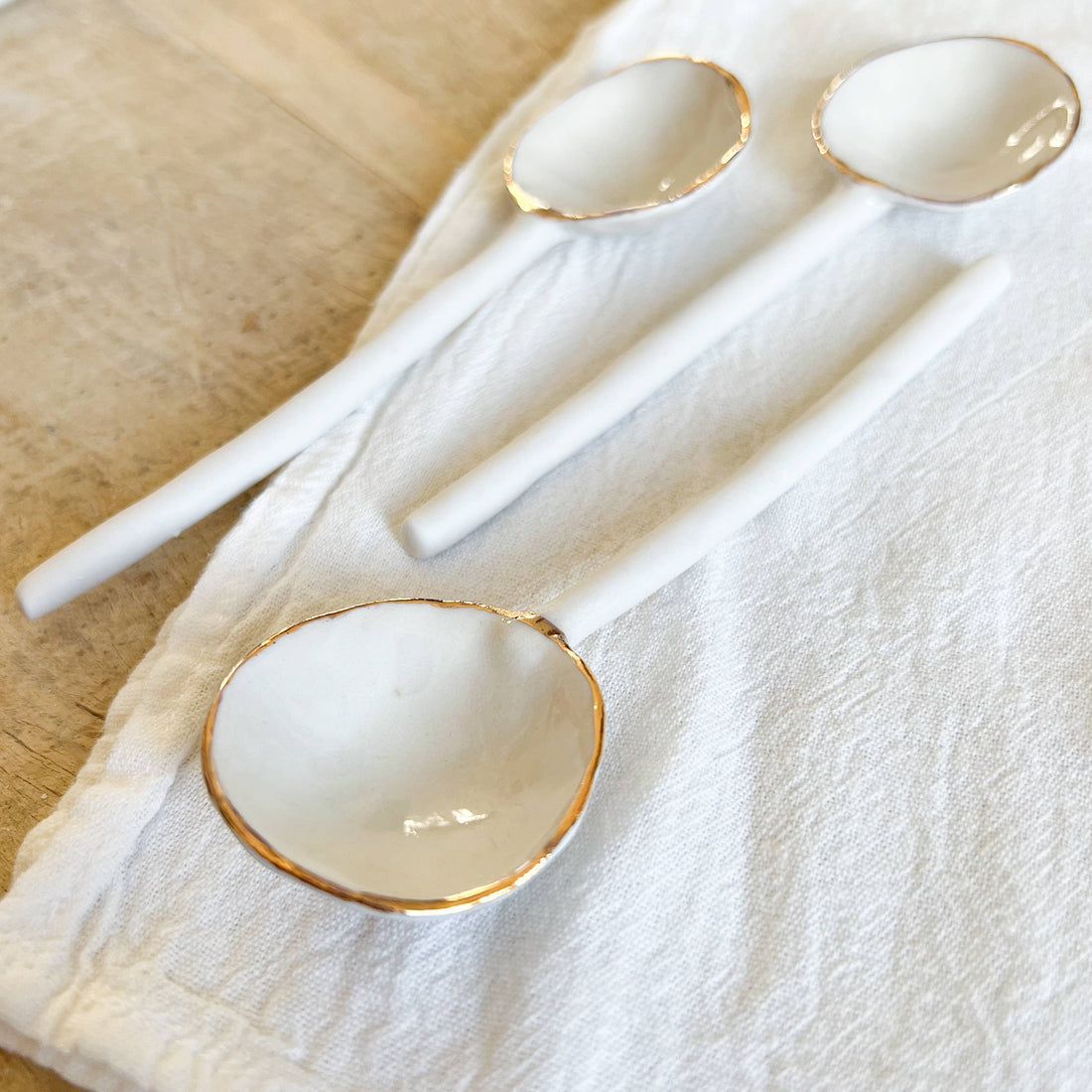 Three Gold-Edged Porcelain Spoons, white porcelain with gold rims, arranged diagonally on a textured white cloth and wood,…
