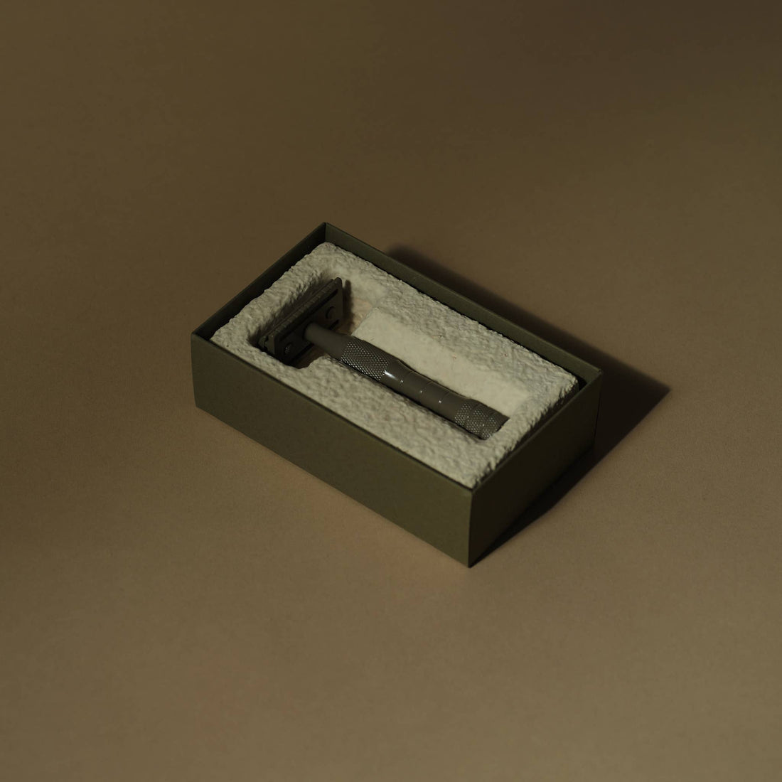 Kelp brass safety razor with textured handle, resting in its open olive-green box with white molded cushioning, angled view.