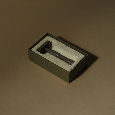
                    
                      Kelp brass safety razor with textured handle, resting in its open olive-green box with white molded cushioning, angled view.
                    
                  