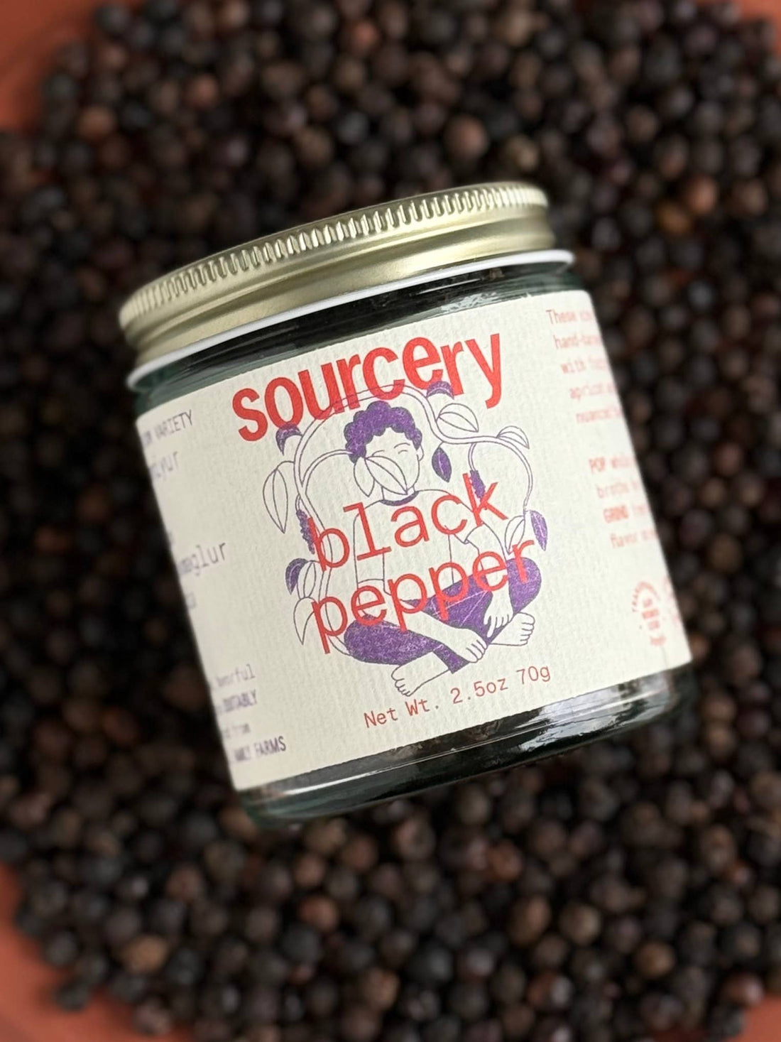 Sourcery Fruity Black Peppercorns spice jar, clear glass with golden lid, colorful illustrated label, tilted overhead view…