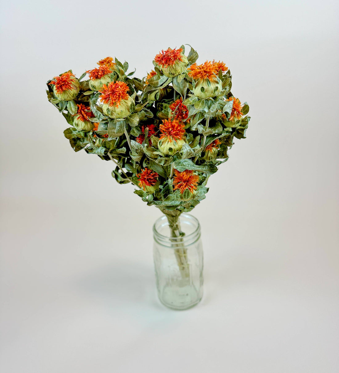 Naturally dried Safflower Orange flowers with green leaves, bundled in a clear glass jar, angled top view.