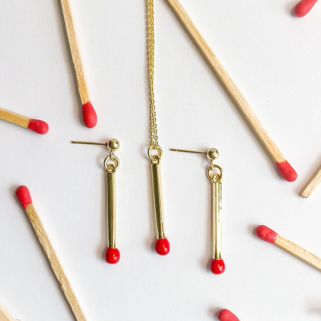 Matchstick necklace and matching drop earrings, gold-plated with red tips, flat lay surrounded by wooden matchsticks for…