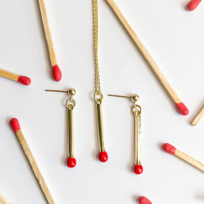 
                    
                      Matchstick necklace and matching drop earrings, gold-plated with red tips, flat lay surrounded by wooden matchsticks for…
                    
                  