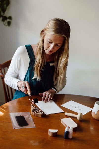 
                    
                      Cross Back Apron, dark teal 100% cotton, worn by woman engaged in block printing activity at a wooden table.
                    
                  
