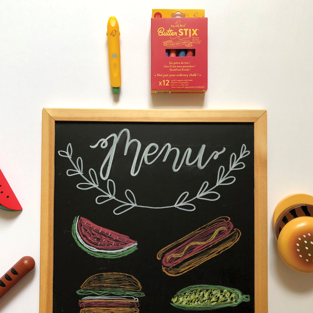 Butterstix Dustless Chalk (12 Pcs) red box & yellow holder, with colorful chalk food drawings on a wooden framed blackboard.