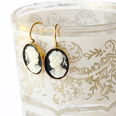 
                    
                      Small Black Cameo drop earrings, white resin figures on black, with gold-plated settings, hanging from the rim of a…
                    
                  