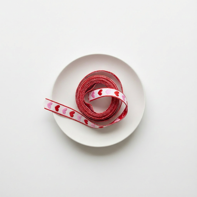 
                    
                      Roll of red ribbon with heart patterns on a white plate against a light gray background
                    
                  