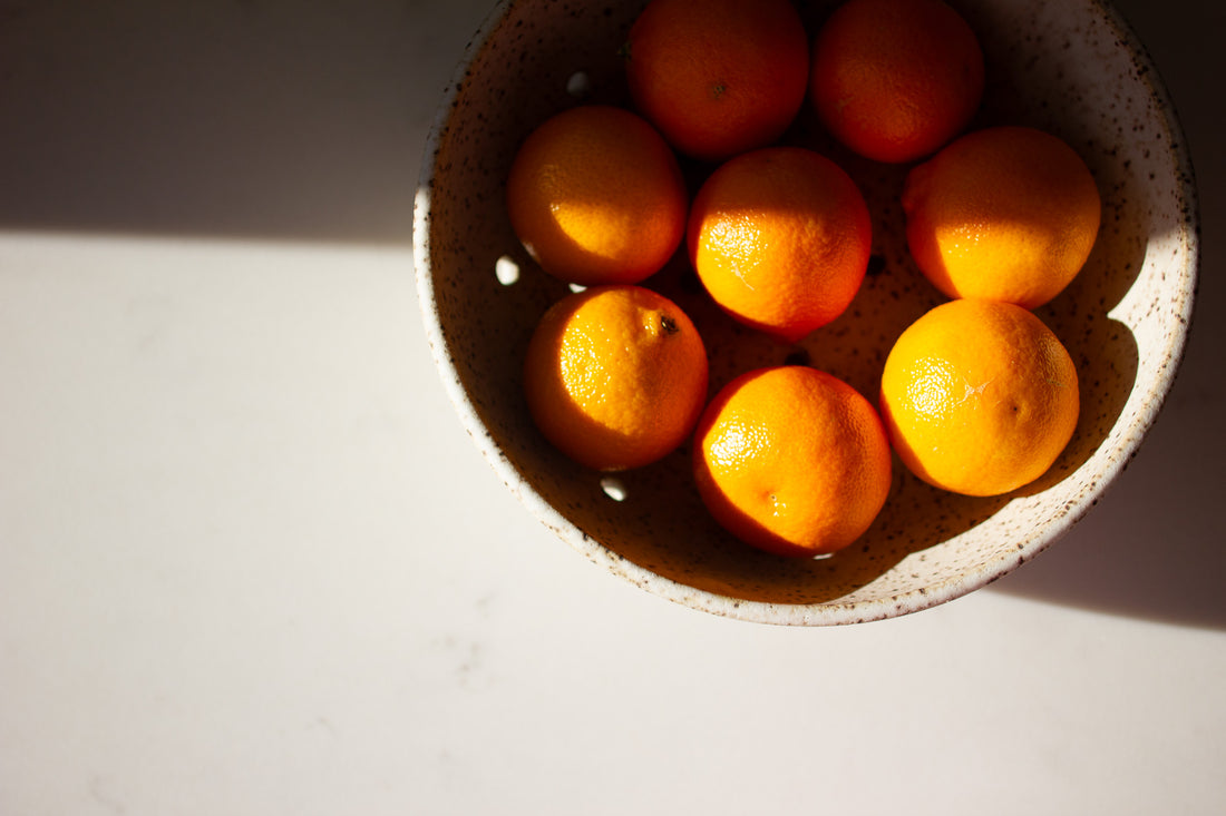 Hand-Thrown Ceramic Colander, speckled natural ceramic, top-down close-up showing textured rim and drain holes, with oranges…