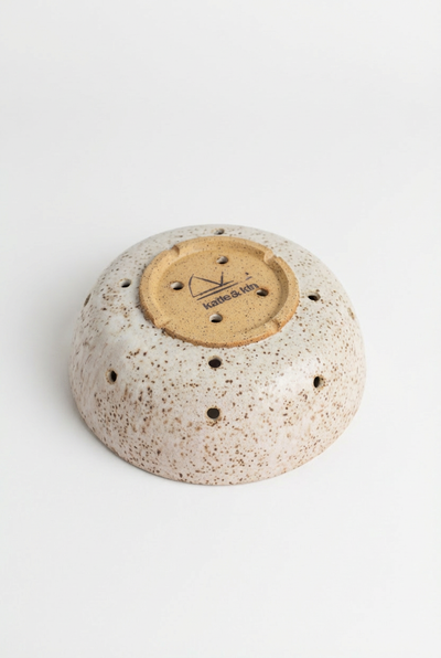 
                    
                      Katie + Kin Hand-Thrown Ceramic Colander, speckled white, underside view showing unglazed earthenware base with brand logo…
                    
                  