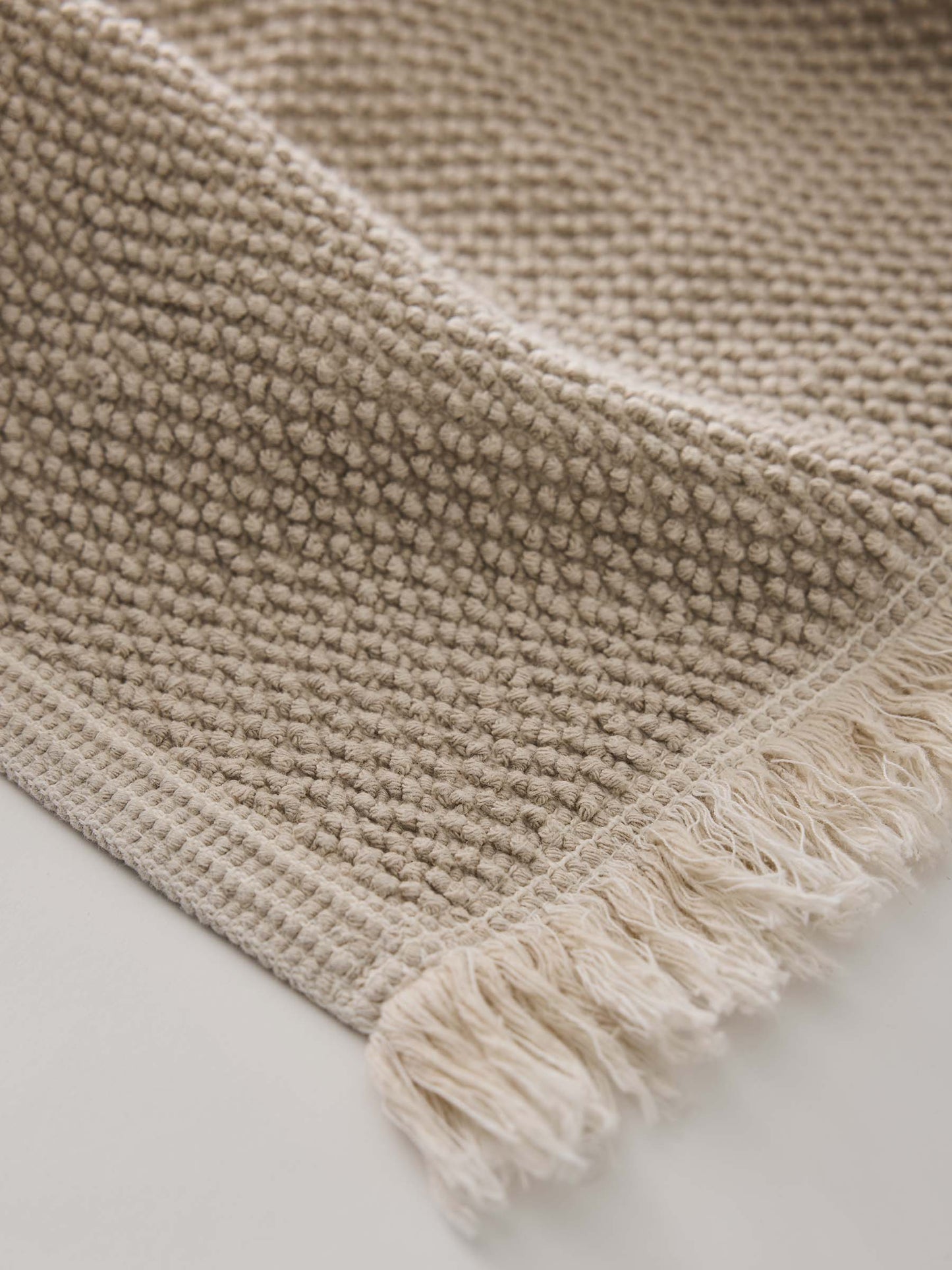 Close-up of a beige woven bathmat with fringes on a light background
