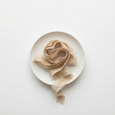 
                    
                      Farmette Plant-Dyed Sari Silk Ribbon Lace, off-white recycled sari silk with frayed edges, coiled on a white plate, flat lay.
                    
                  