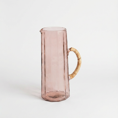 
                    
                      Pink handblown hammered glass pitcher, visible vertical ridges, and a natural rattan-wrapped handle.
                    
                  