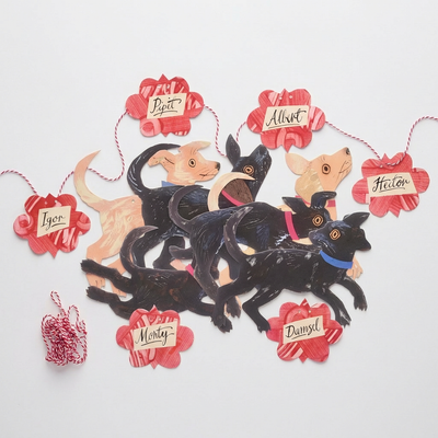 
                    
                      Puppy Parade Garland, Mark Hearld illustrated paper cutouts of black and tan dogs, red flower name tags, flat lay detail.
                    
                  